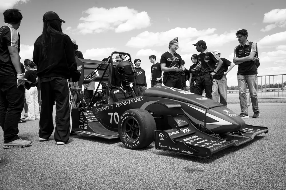 Black and white photo of team members gathered around the car at the track